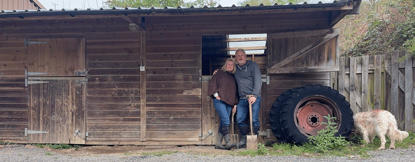 Two people standing in front of a wooden shed with a large tire and a dog nearby.